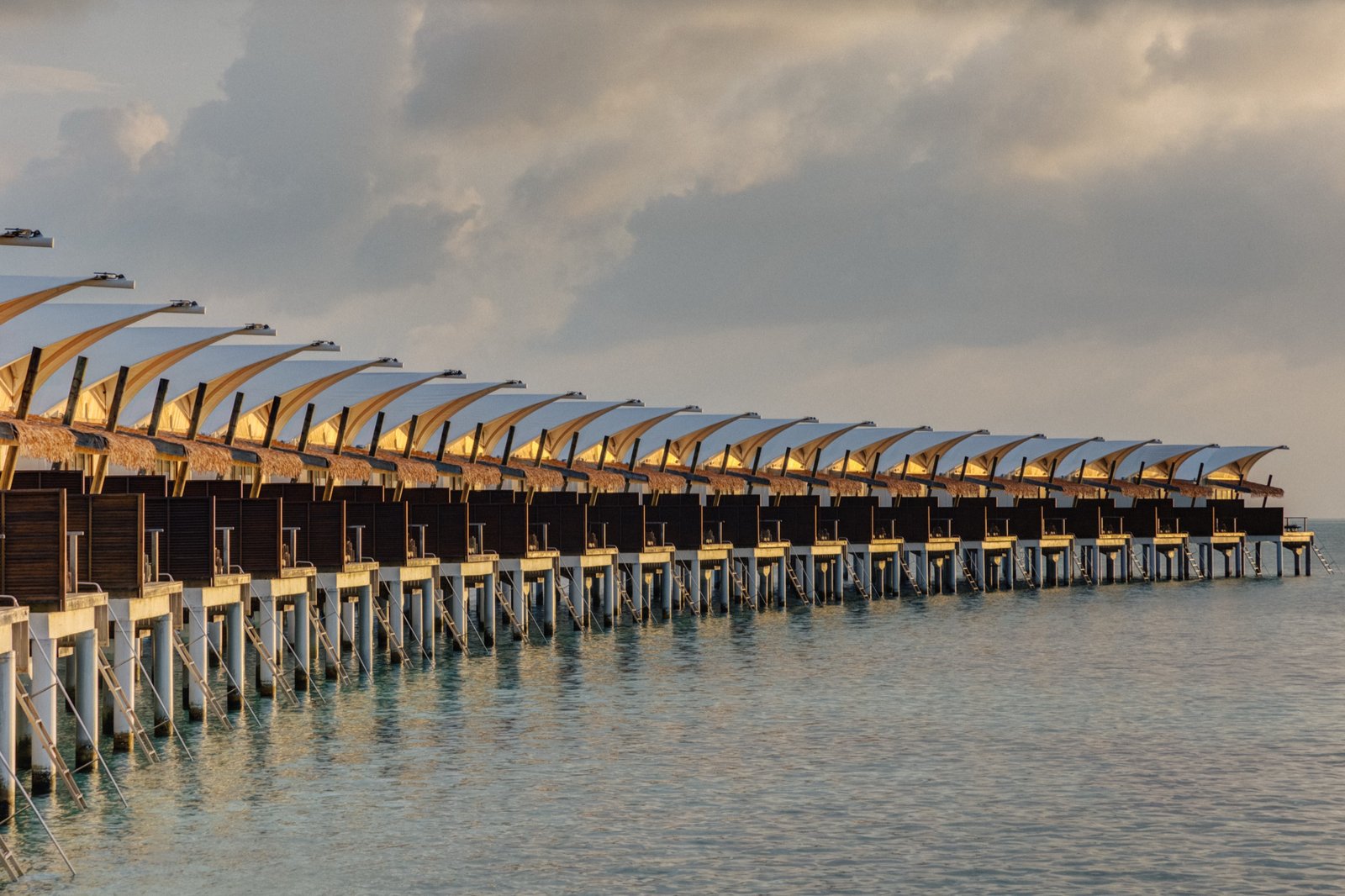 Overwater bungalow row at golden hour, Cinnamon Hakuraa Huraa Maldives — the Sunset Water Bungalow shares this same 70 m² overwater design on the sunset-facing arm