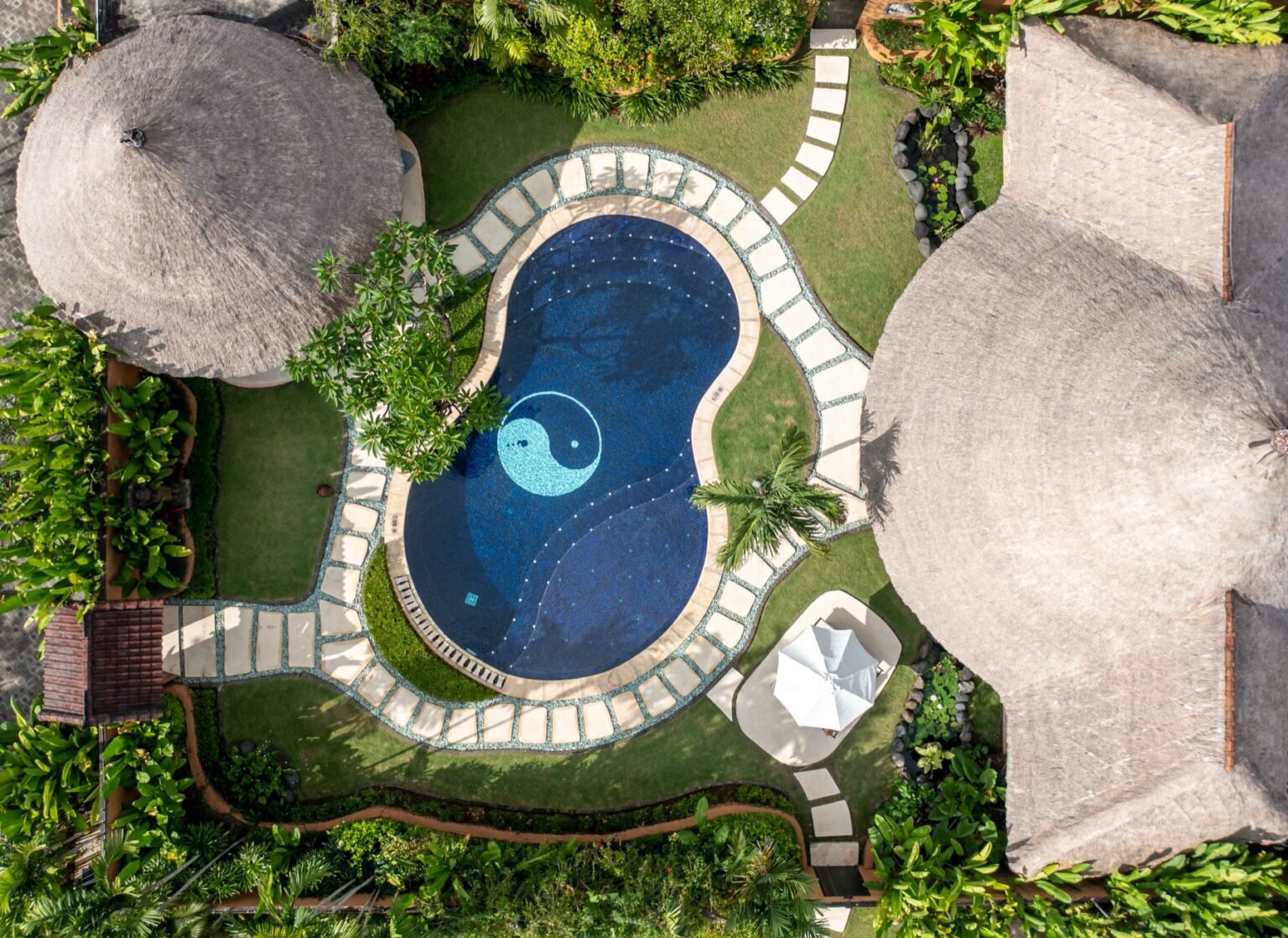 An aerial of a villa with a curved pool, a thatched pavilion and lawn inside its garden walls.