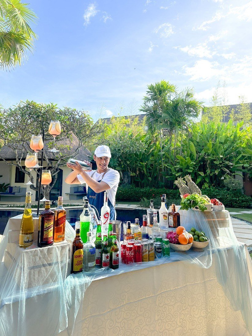 A bartender shaking a drink behind a cloth-covered table of bottles, mixers and fruit set up beside a villa pool.
