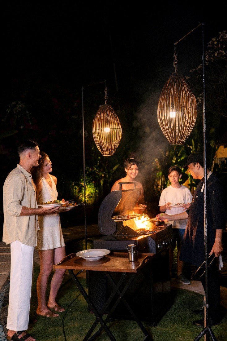 A chef grilling at an outdoor barbecue after dark under two woven lanterns, with guests standing alongside holding plates.
