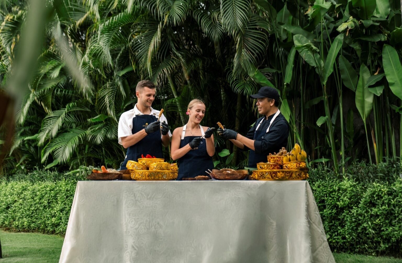 Three people at a cloth-covered table on a lawn, with Balinese ingredients and carved fruit laid out in front of palms.