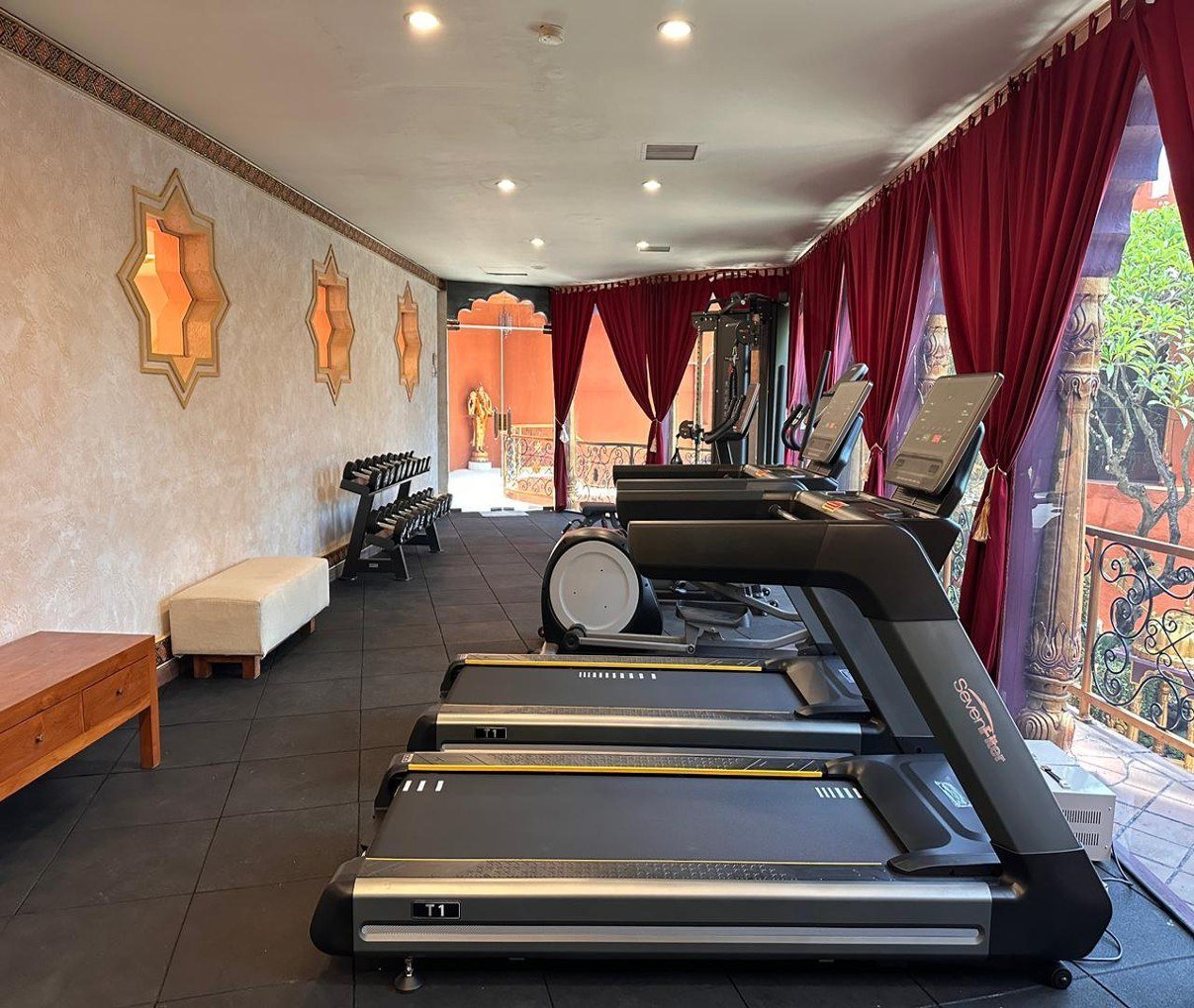 A row of treadmills and a cross-trainer in a long room with patterned wall niches, red drapes and a view to a balcony.