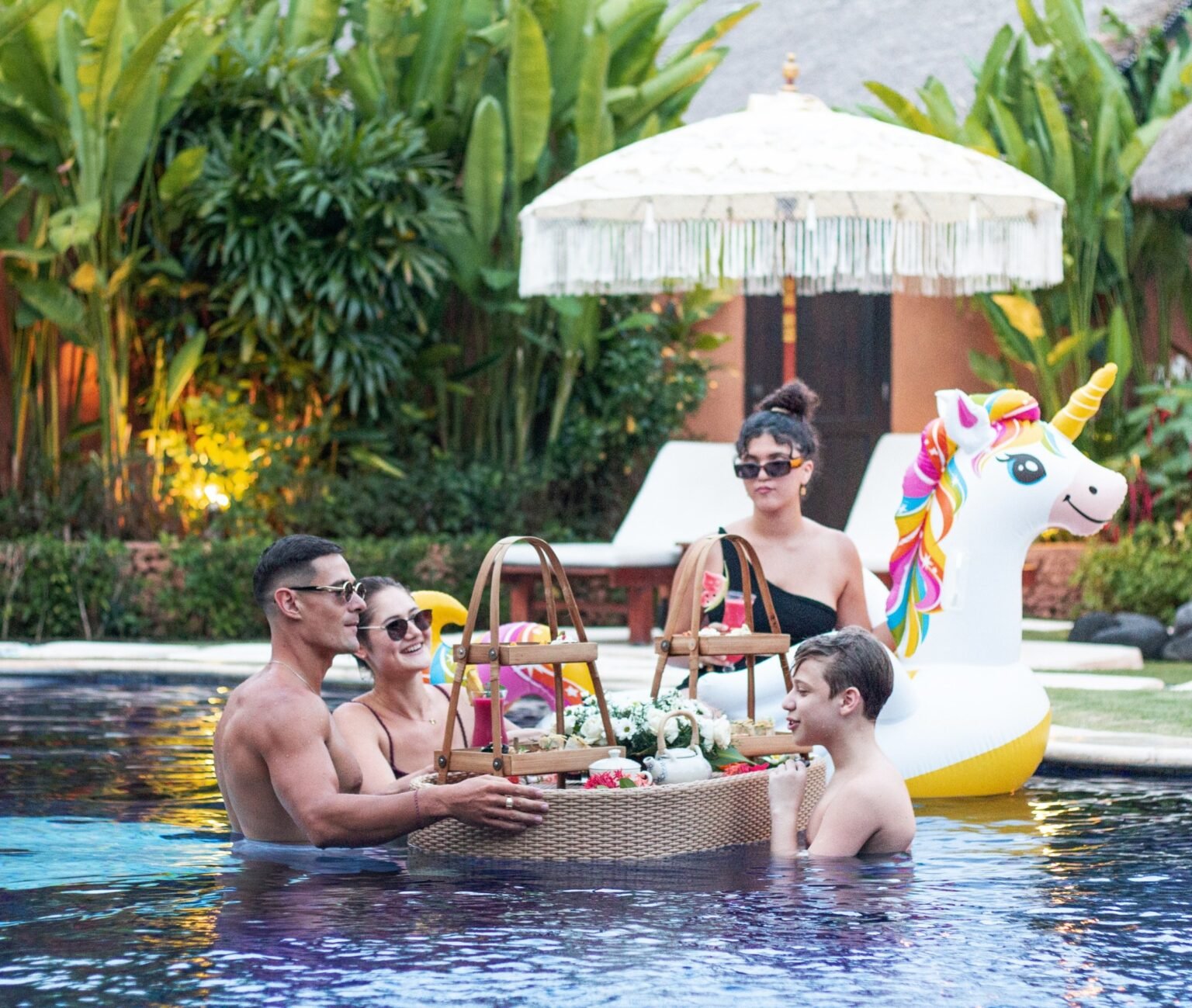 A family in a villa pool around a floating tray of food, with a fringed parasol behind them and an inflatable unicorn alongside.