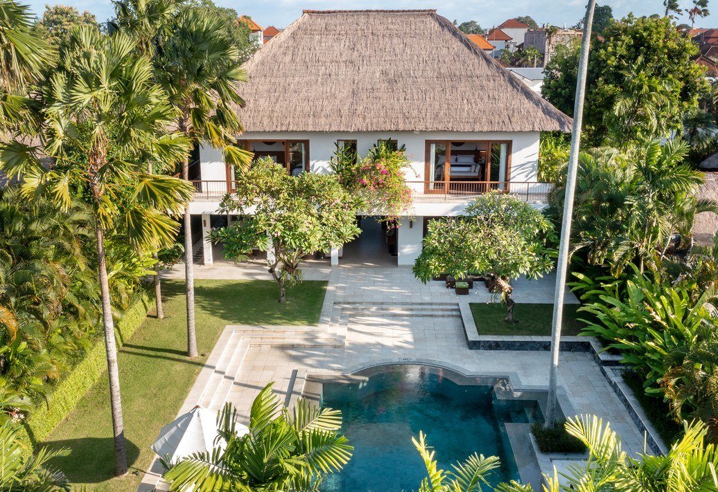 A two-storey villa with a thatched roof seen across its garden, with a pool and paved terrace below.
