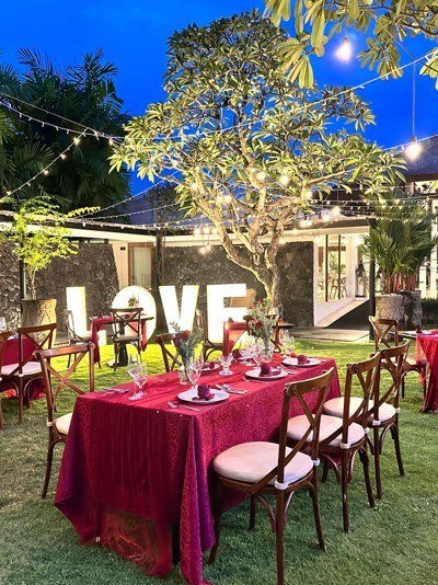 A long table dressed in red and set for dinner on a lawn at dusk, with lit letters spelling LOVE and a tree strung with lights behind.