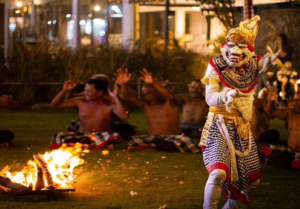 A masked Balinese dancer in costume in front of a seated chorus around a fire after dark.