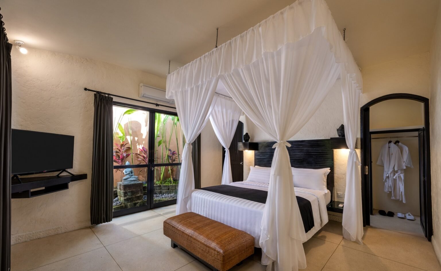 A villa bedroom with a four-poster bed hung with white netting, a wall-mounted television and glass doors to the garden.