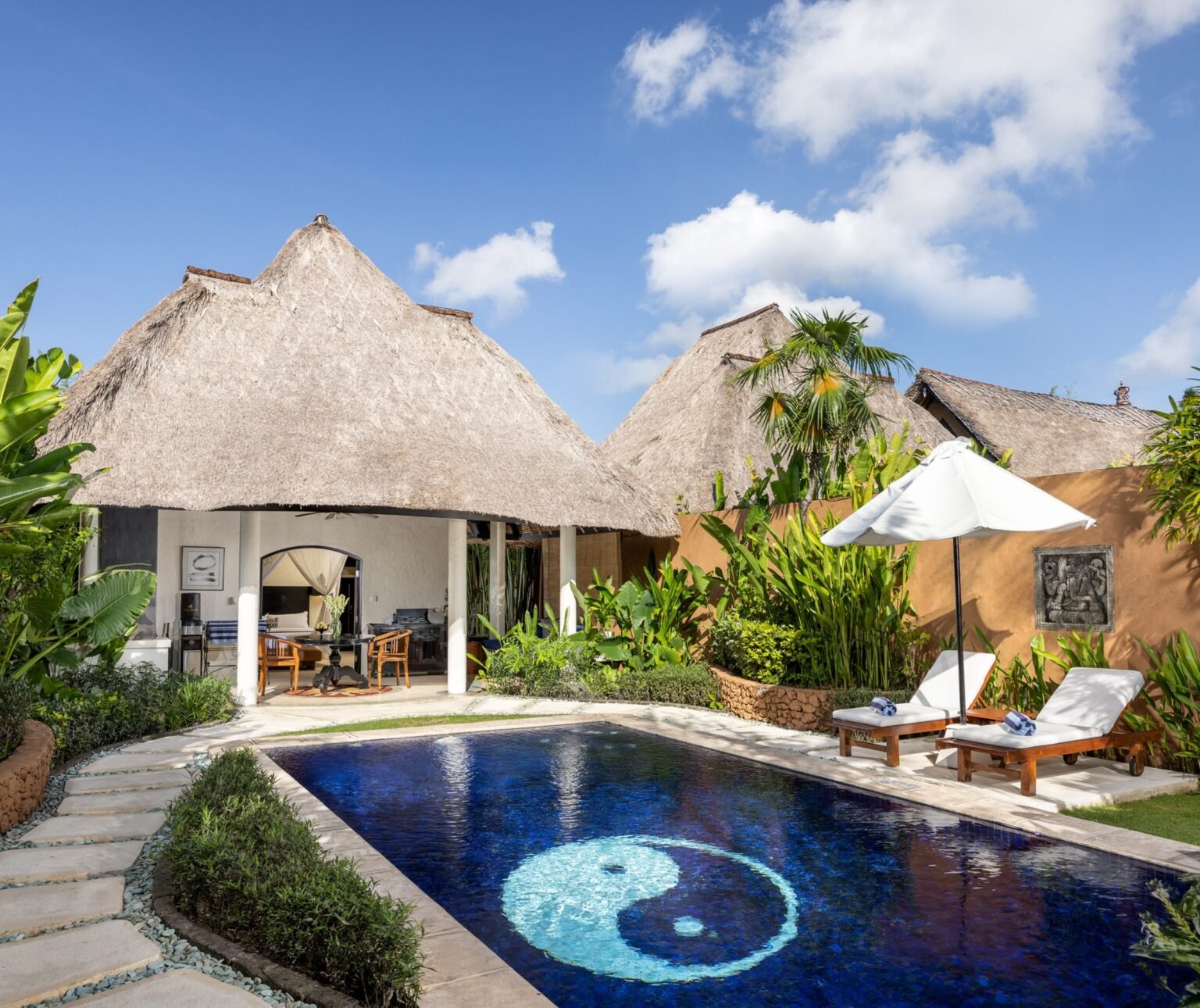 A villa with two thatched roofs beside its own pool, the pool floor patterned with a mosaic yin-yang, with sun loungers and a parasol on the paving.