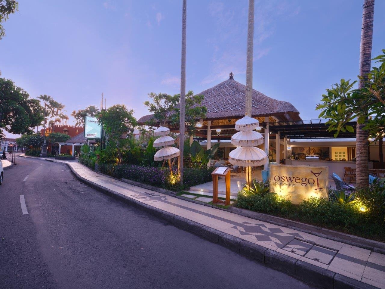 The restaurant seen from the street at dusk, with fringed parasols, a thatched roof and the outlet's own sign lit beside the path.