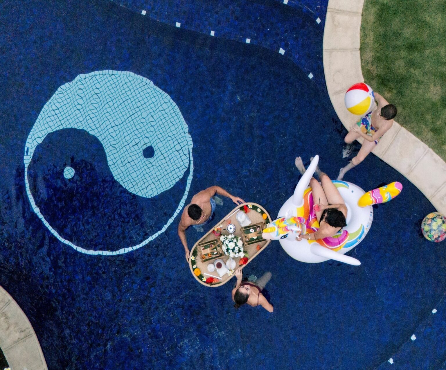 An overhead view of a villa pool with a mosaic yin-yang floor, a floating tray of food and swimmers around it.