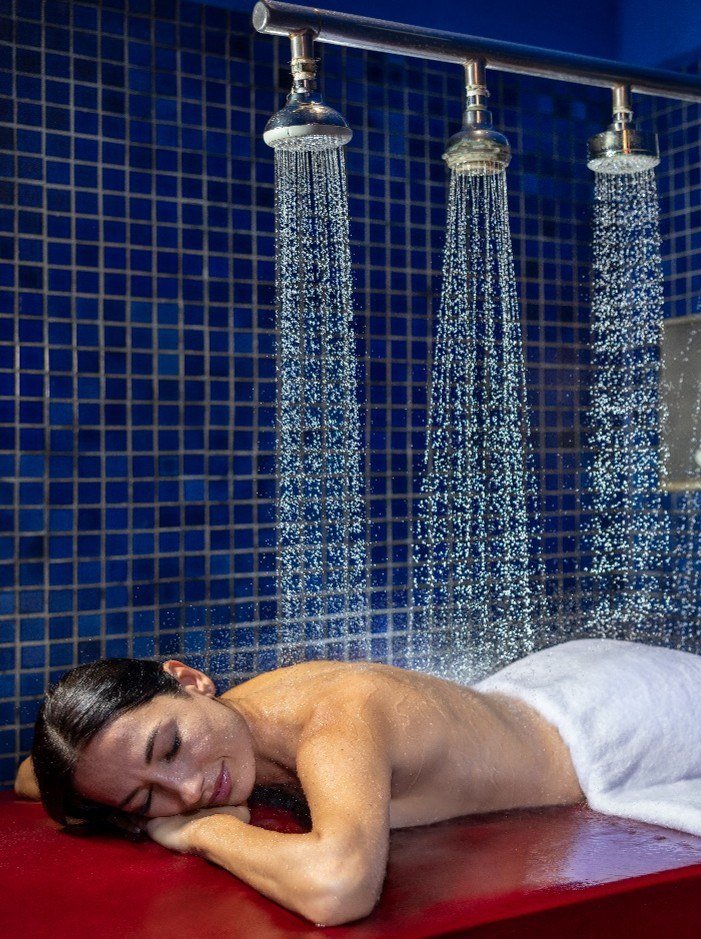 A person lying face down on a treatment bed beneath a row of overhead shower heads in a blue-tiled room.