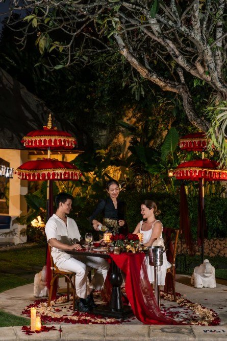 A table set for two on a lawn after dark between red ceremonial parasols, with a member of staff pouring a drink.