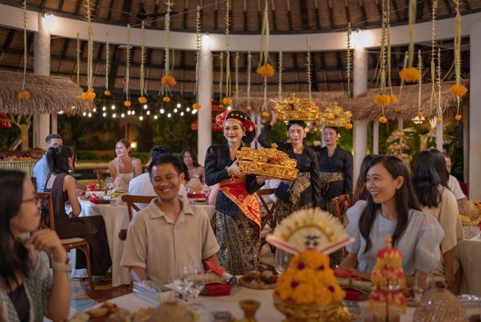 Staff in Balinese dress carrying tiered offerings into a round pavilion hung with lanterns, where guests sit at dressed tables.