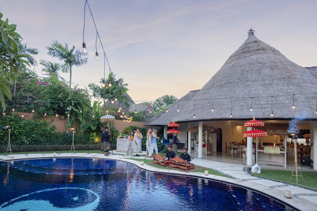 A villa pool at dusk with strings of lights overhead, red ceremonial parasols, musicians seated on the paving and a thatched pavilion behind.