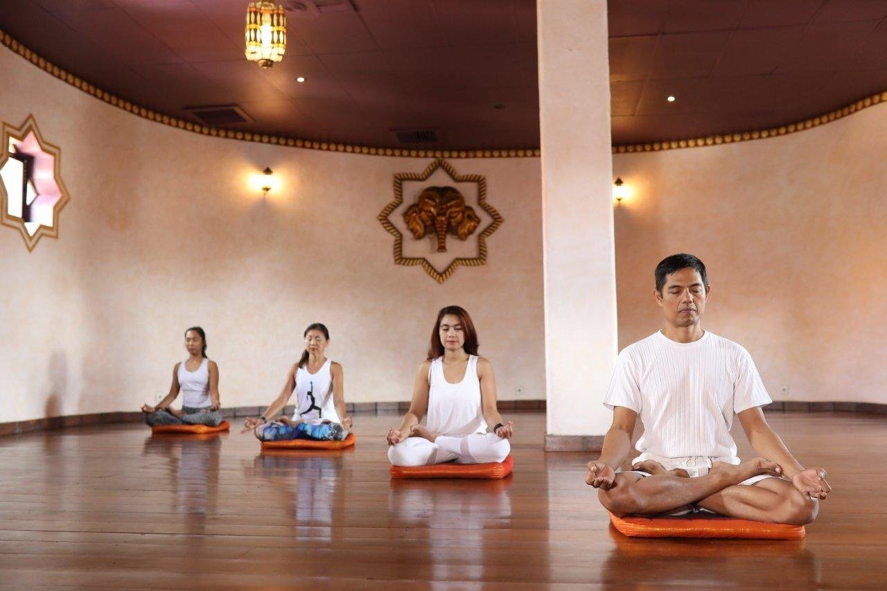 Four people sitting cross-legged on cushions on a timber floor in a round room with a carved wall panel behind them.