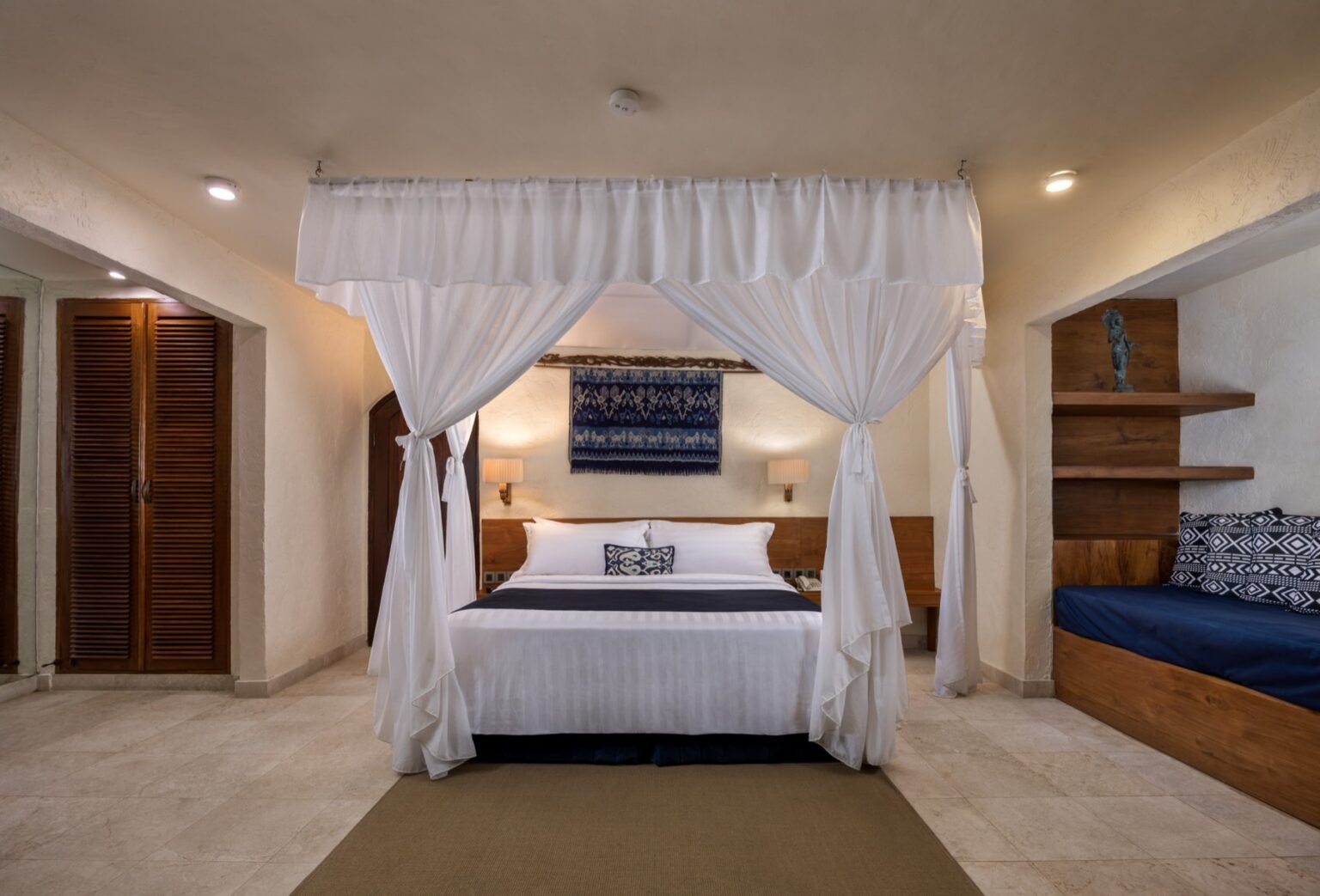 A villa bedroom with a four-poster bed hung with white drapes, and built-in shelving beside it.