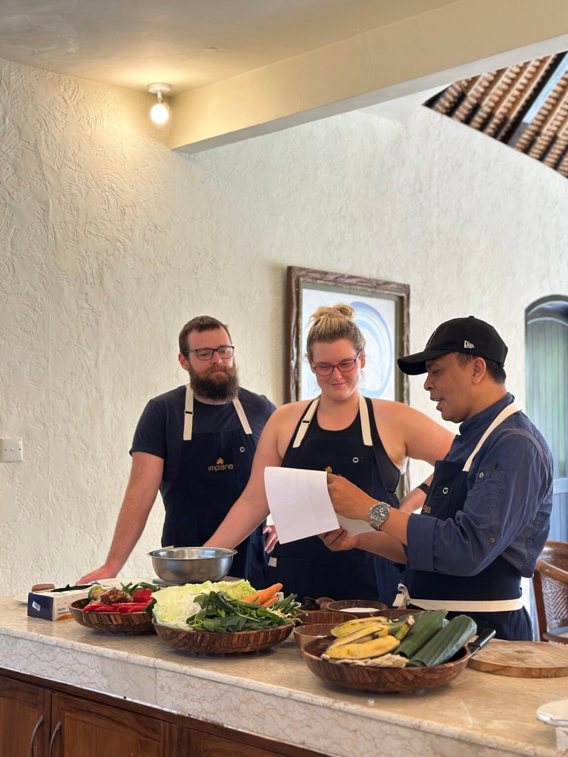 Three people in aprons standing together over a recipe sheet during a cooking class.