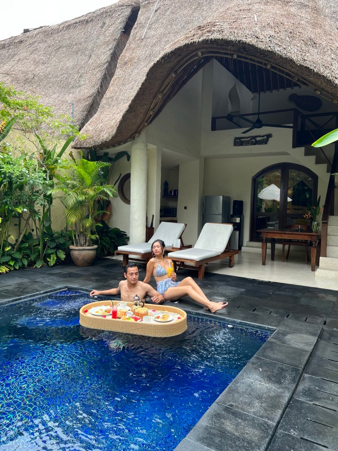 Two people sitting at the edge of a villa pool with a floating breakfast tray in the water beside them.