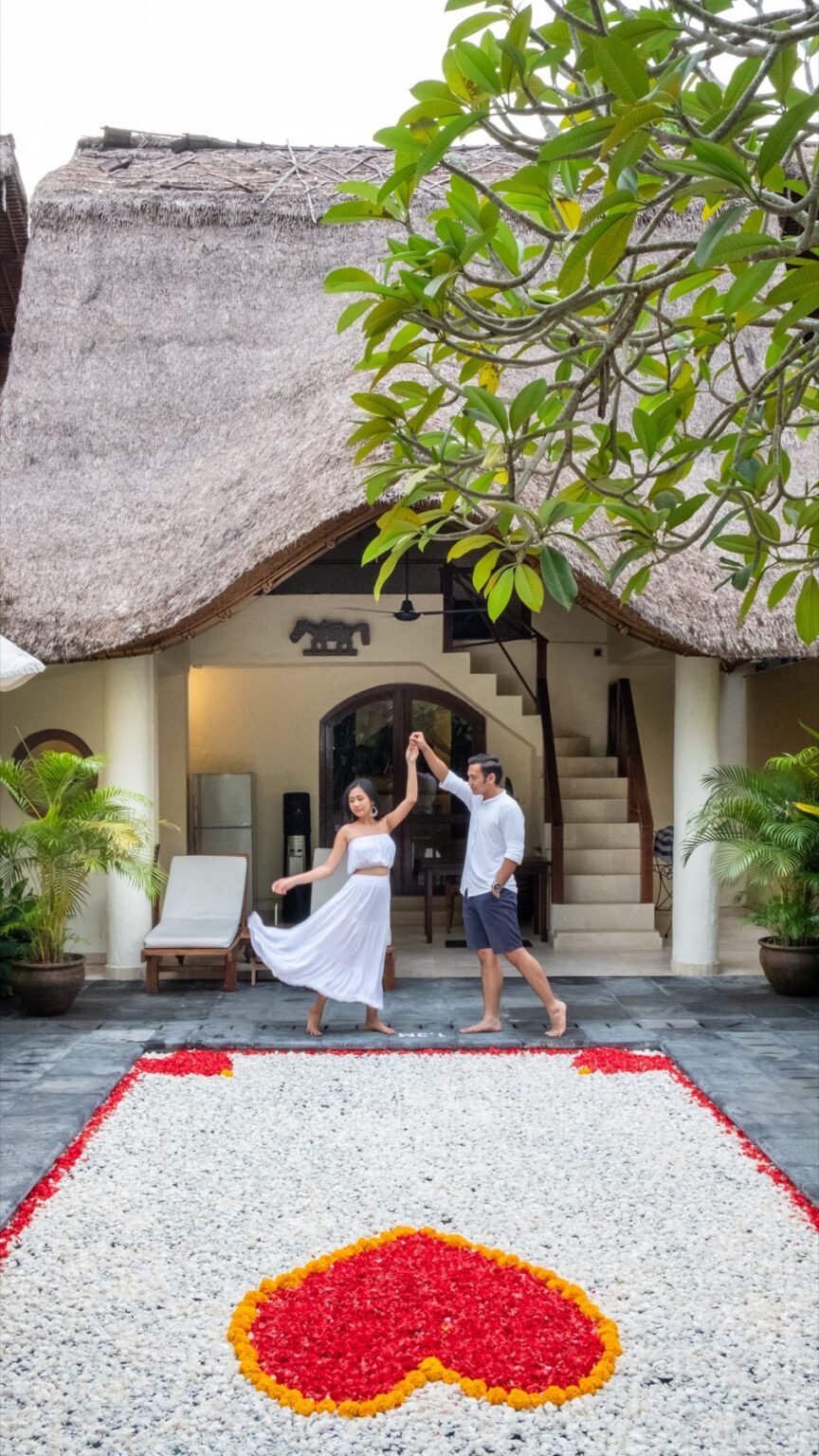 Two people dancing on a villa terrace beside the pool, under a thatched roof.