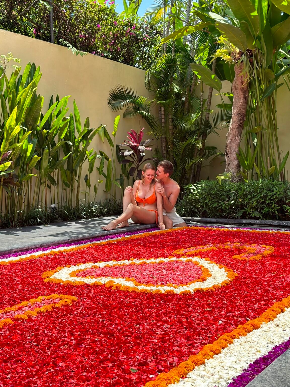 Two people sitting at the edge of a pool whose surface is covered in red petals arranged in a heart.