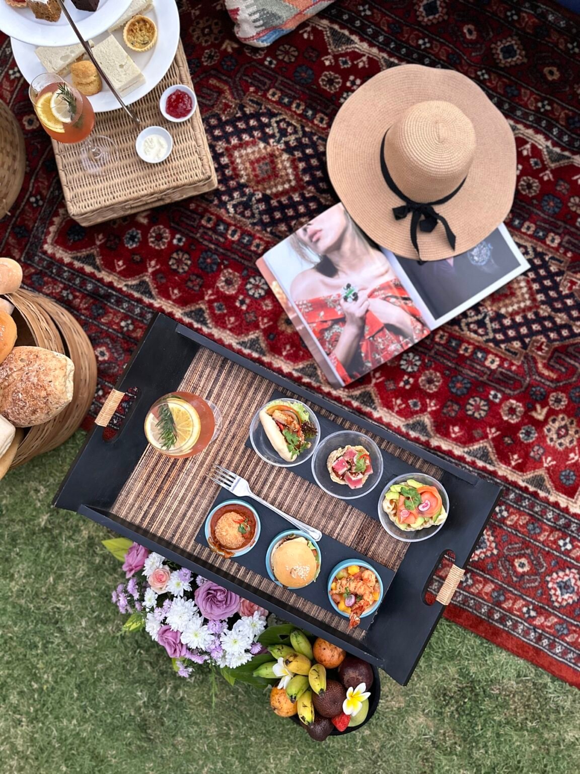 An overhead view of a picnic laid out on a rug: dishes, a basket and cushions.