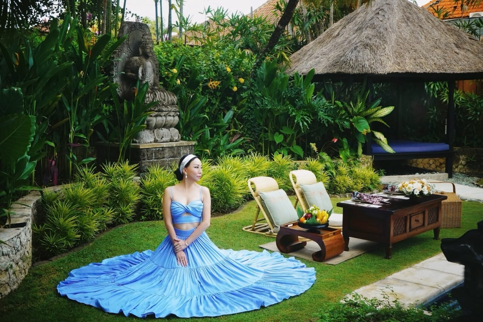 A woman in a long blue gown on a lawn beside a thatched pavilion and loungers.