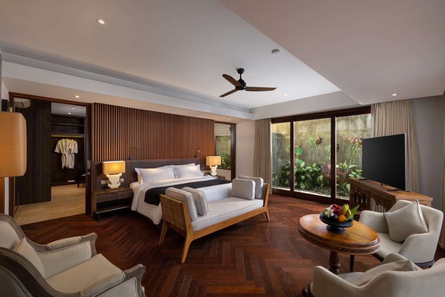 A villa bedroom with a king bed, a ceiling fan and an armchair by the window.