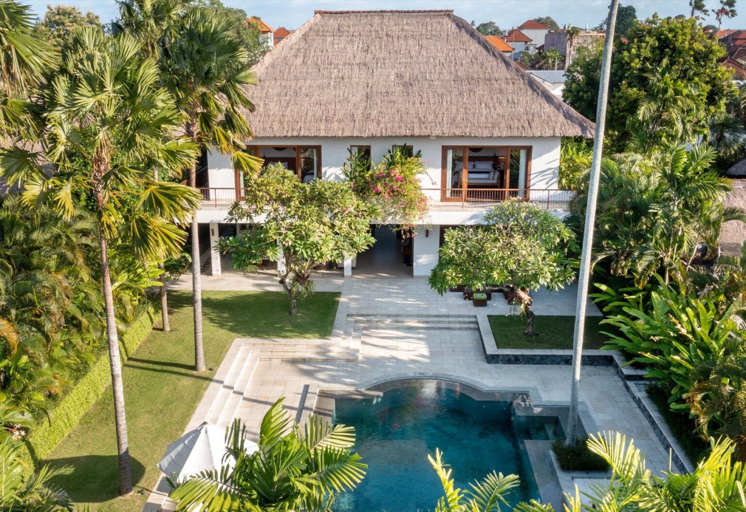 A villa exterior with a thatched roof seen across its pool and lawn.