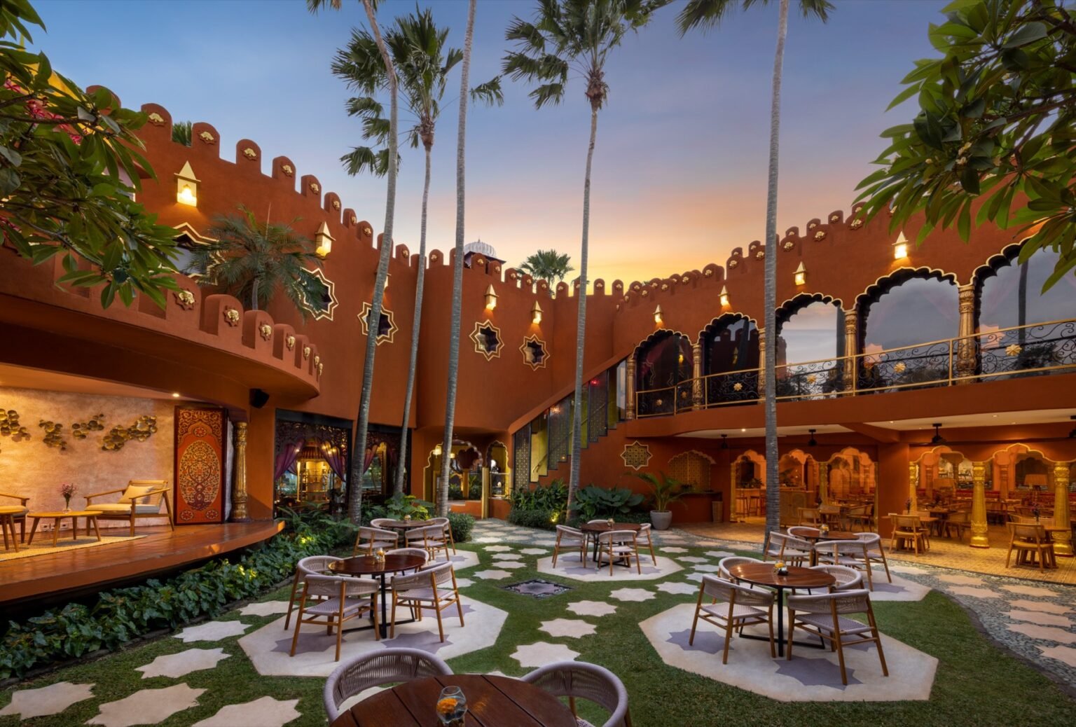 A terracotta building with scalloped arches and star-shaped lattice windows at dusk, with dining tables set on the lawn in the courtyard and palms overhead.