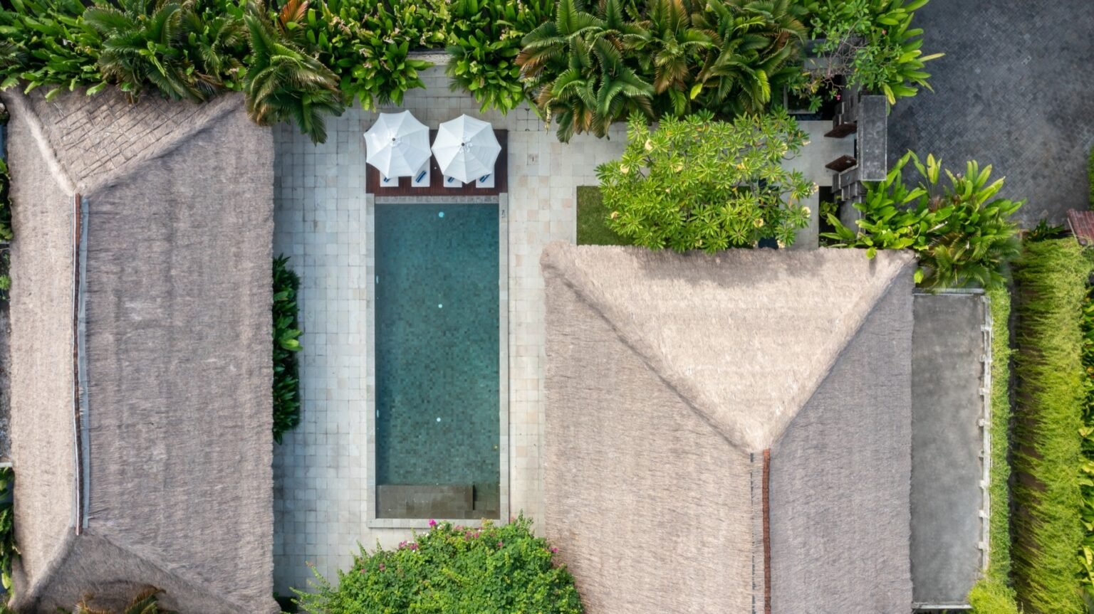 An overhead view of a villa, its pool and its lawn, with thatched roofs on two sides.