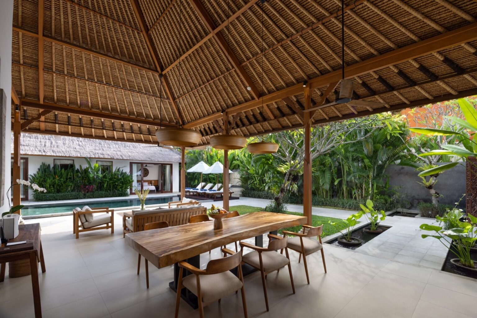 An open-air dining table under a thatched roof, looking out to the pool and loungers.