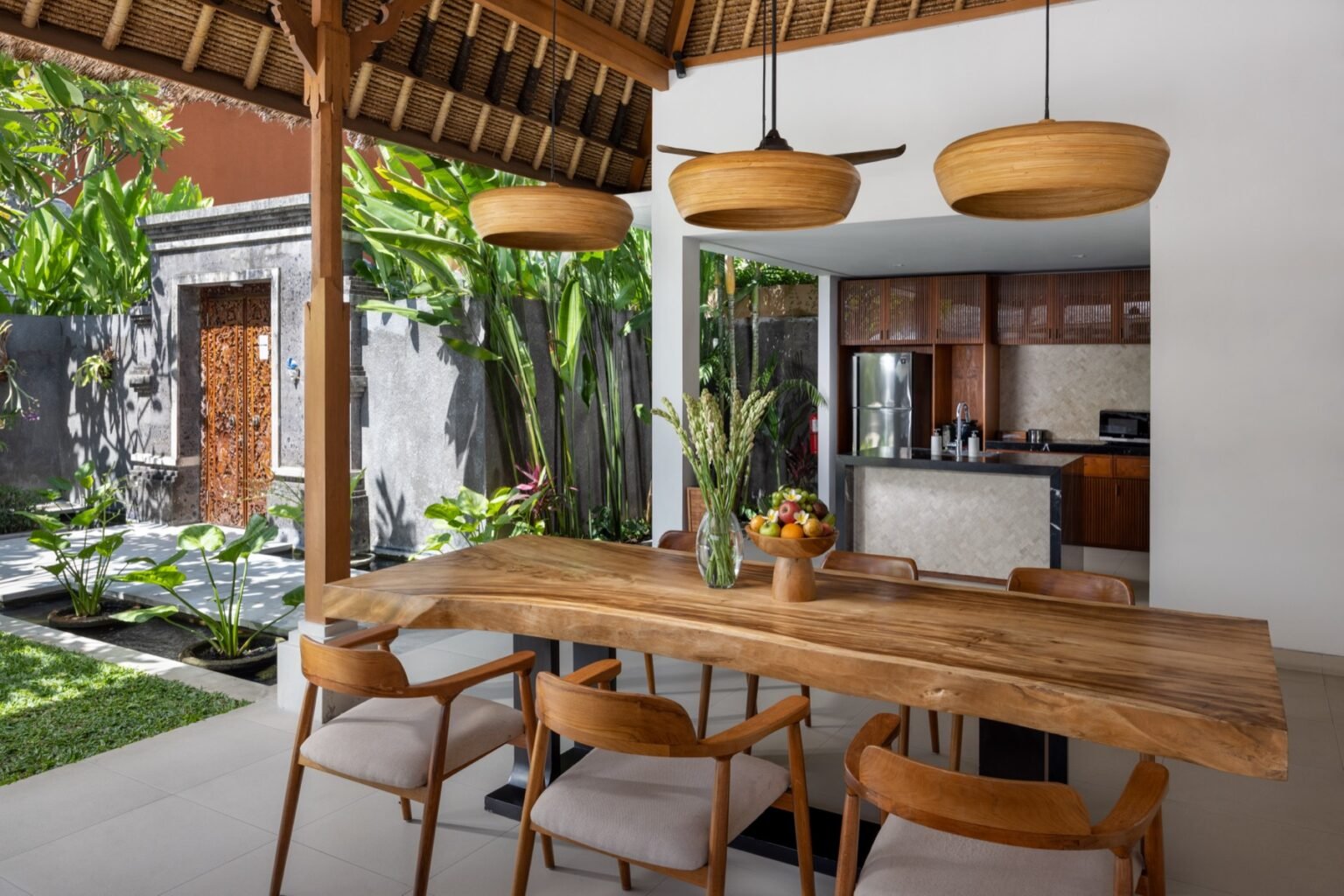 A long dining table with pendant lights above it, beside an open kitchen and a garden.