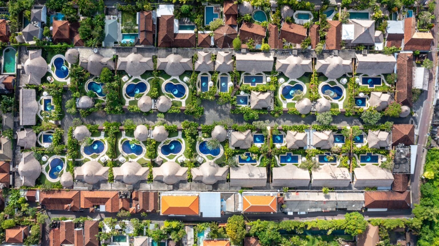 An overhead view of thatched villa roofs in rows, each with its own blue pool and garden.