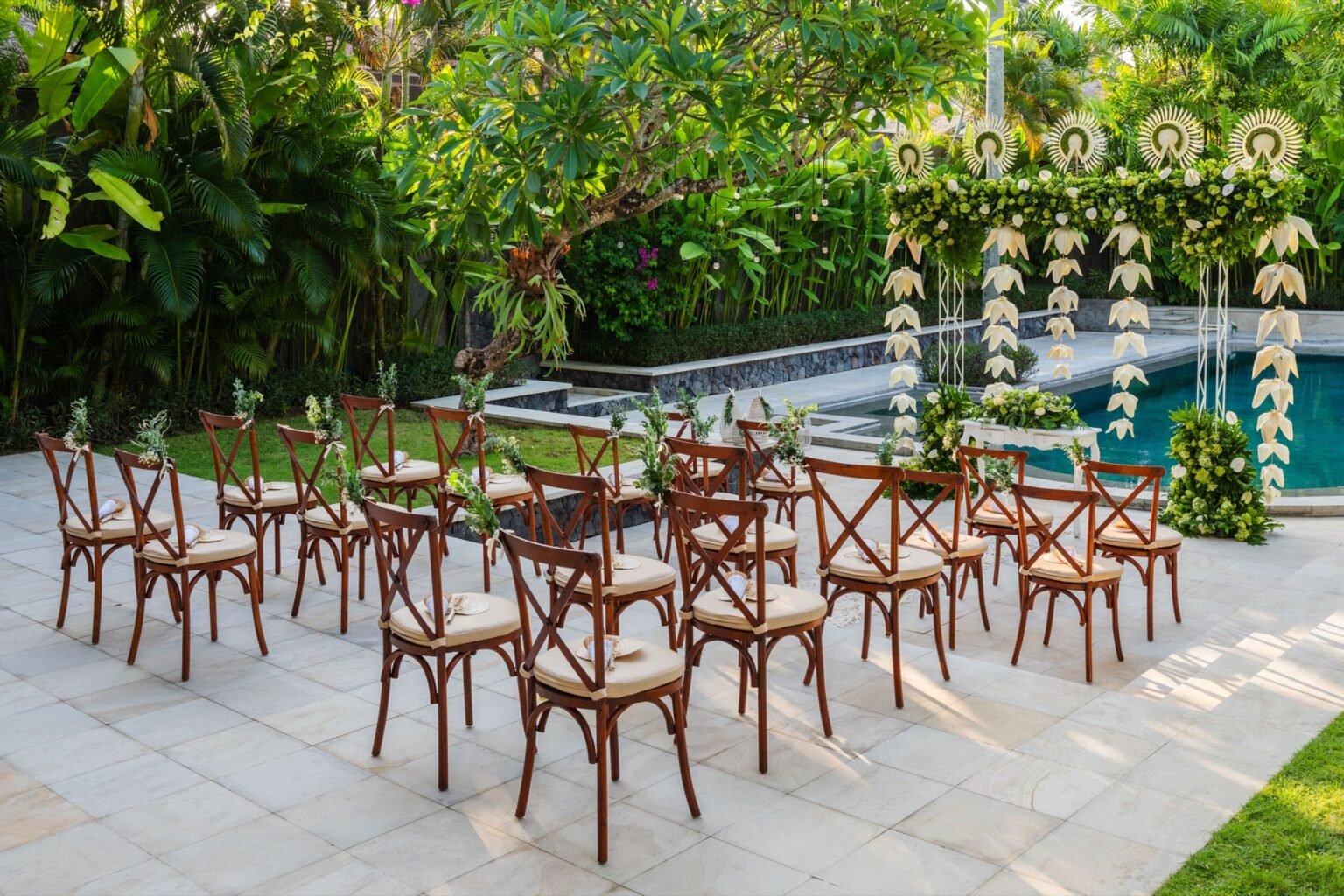 Rows of wooden chairs set out beside a villa pool facing a floral arch.
