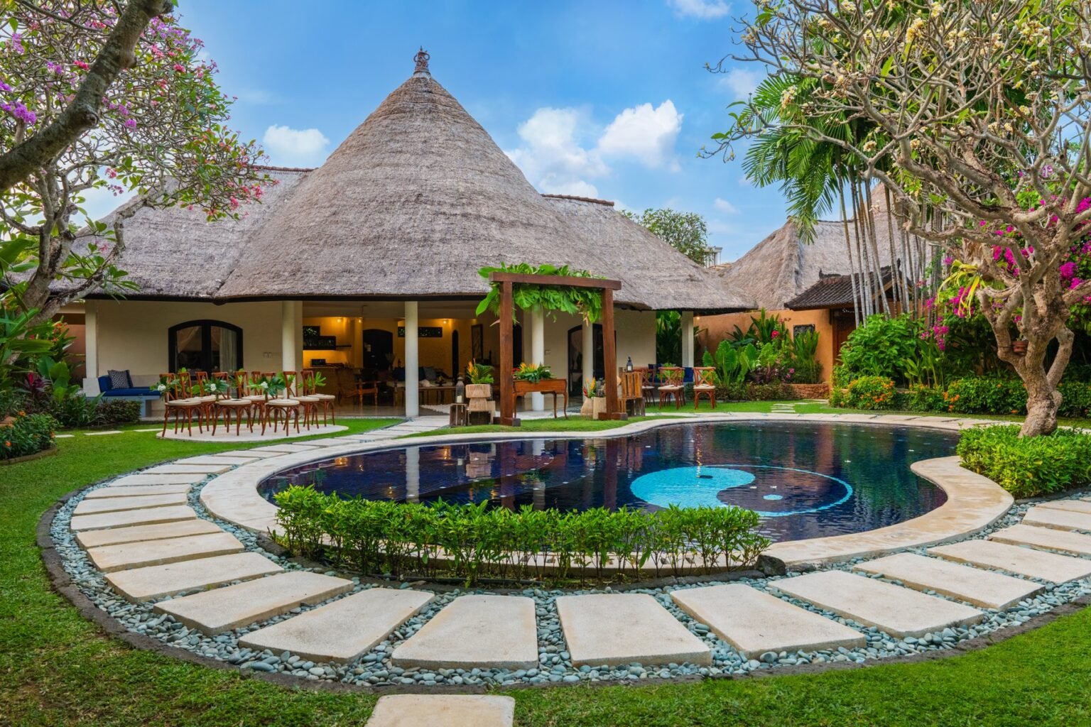 A villa with a thatched roof seen across its pool and lawn, set up for an event.