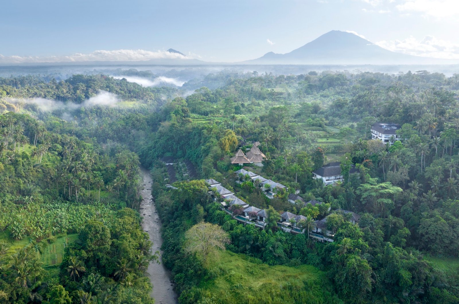 An aerial view of the Ayung river gorge in morning mist, with villas stepping down a jungle hillside and a volcano on the horizon