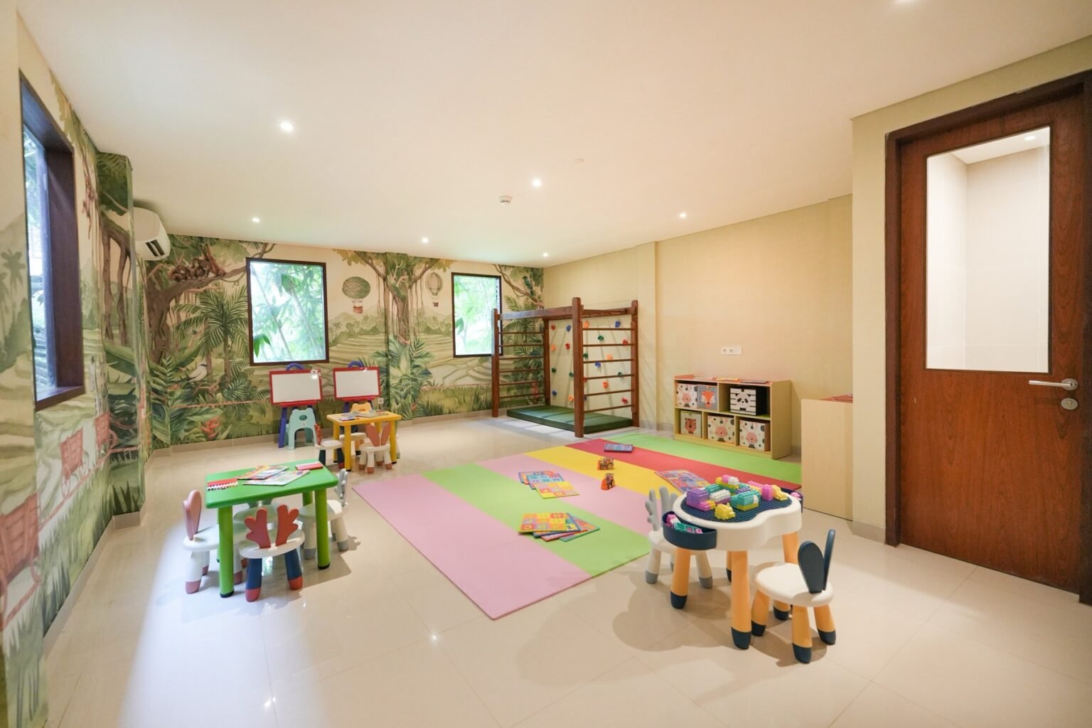 A children's playroom with a painted jungle mural, small tables and chairs, soft floor mats and a climbing frame.