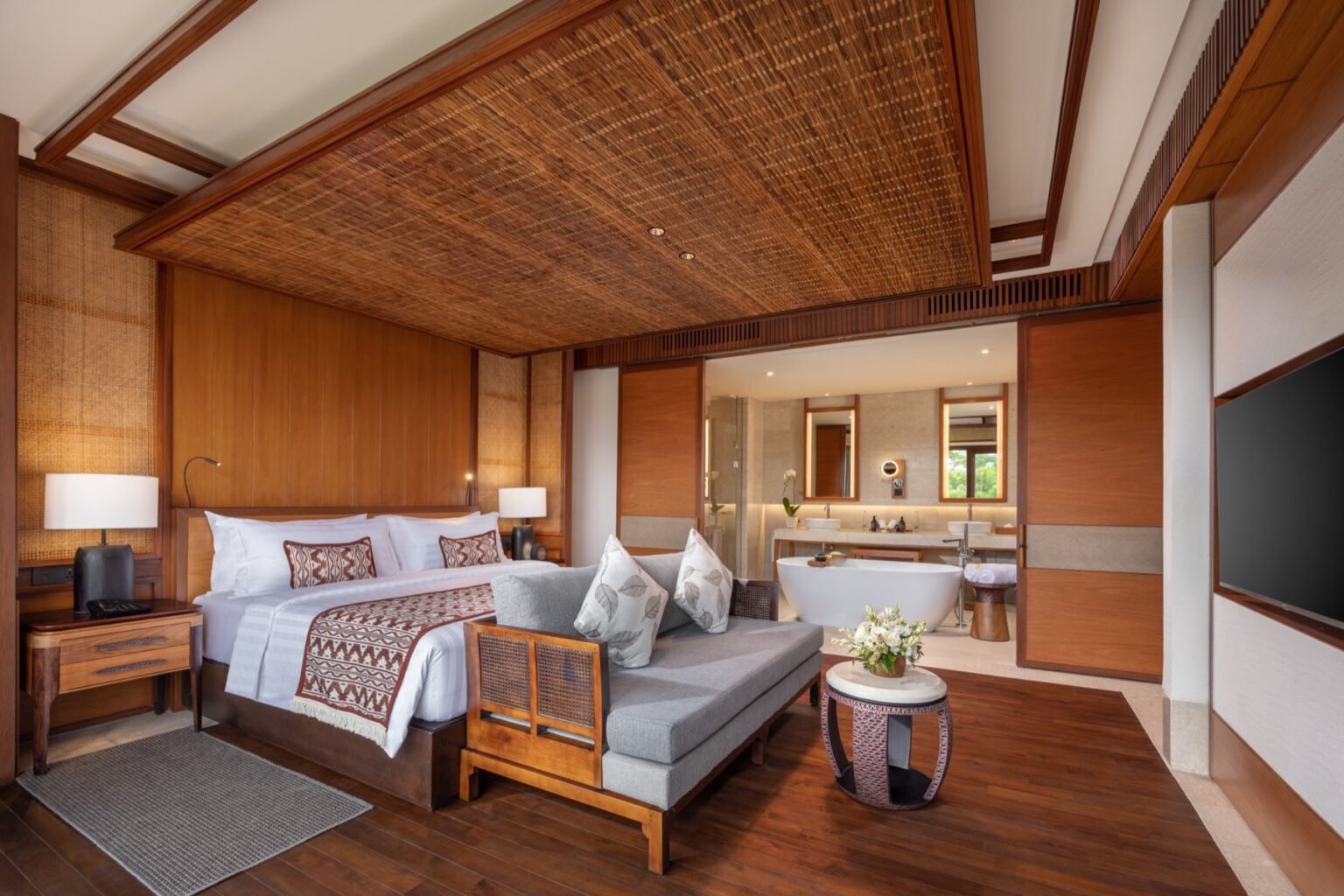 A suite bedroom with a bed, a sofa and a seating area under a timber ceiling.