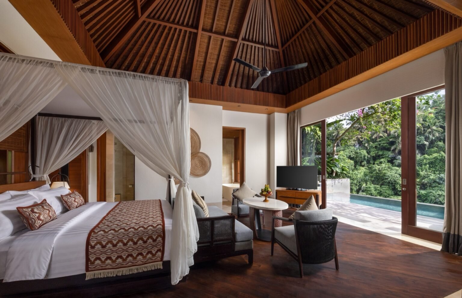 A villa bedroom with a four-poster bed and mosquito net under a peaked timber ceiling, with open doors onto a private pool and jungle beyond.
