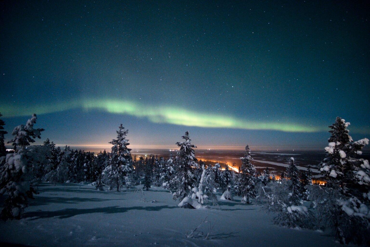 Northern lights over the snow-covered fell forest, with the valley lights below — Levin Iglut – Golden Crown, Levi