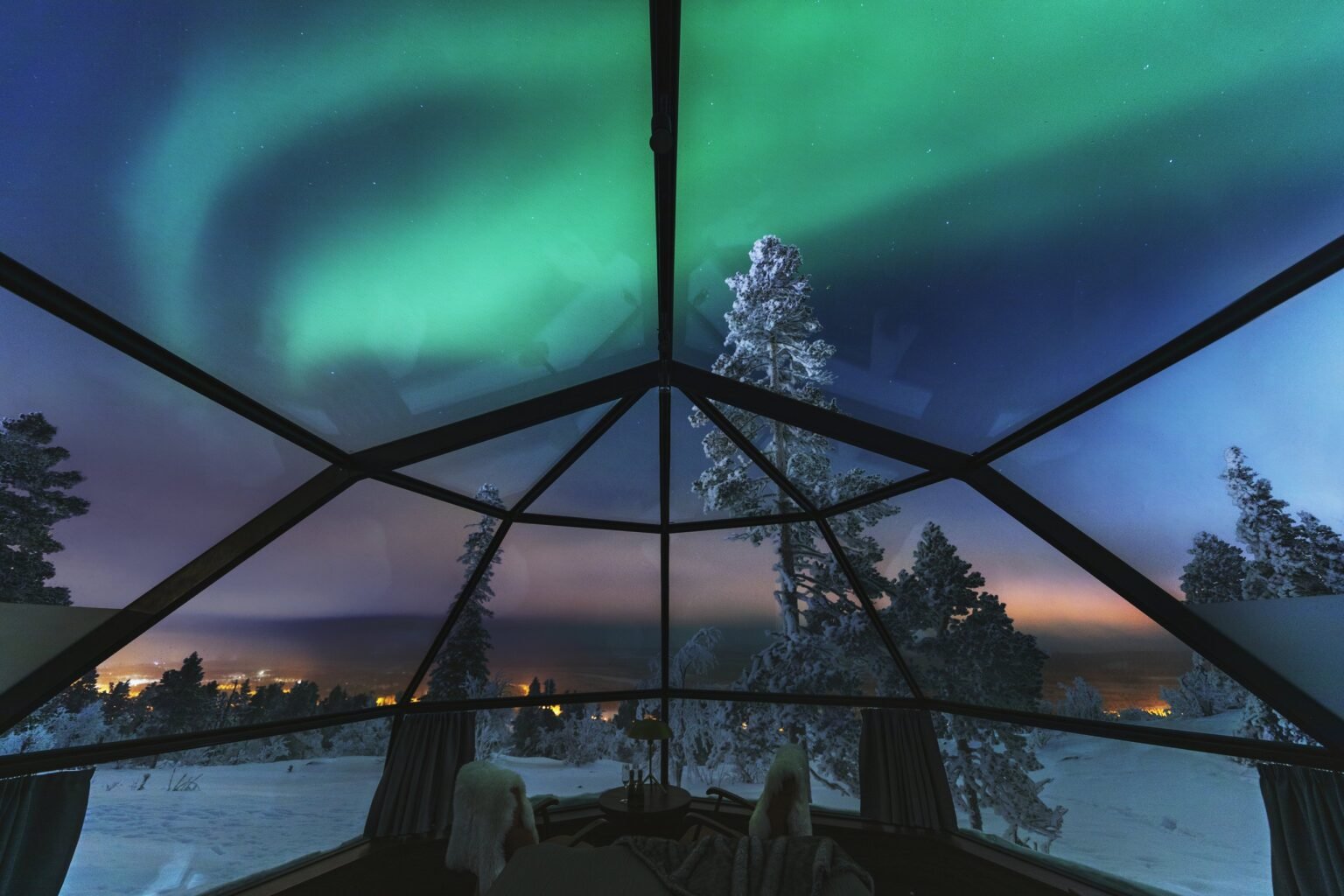 Aurora through the glass roof, seen from inside an igloo, with snow-covered pines and the valley lights beyond — Levin Iglut – Golden Crown, Levi