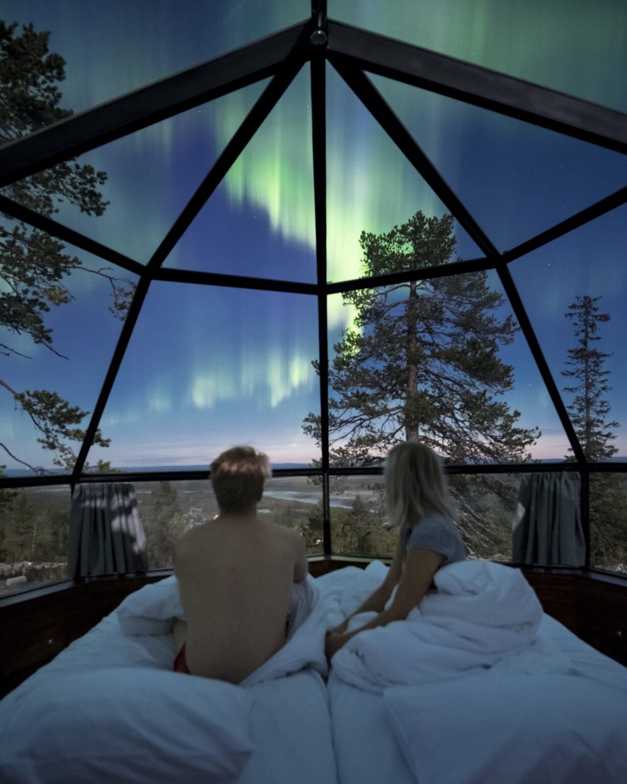 Two guests on the bed looking out at the aurora through the glass roof of an igloo — Levin Iglut – Golden Crown, Levi