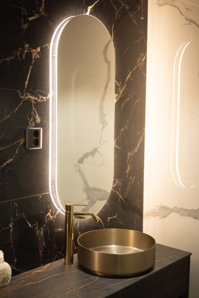 A brass basin below a lit oval mirror against a dark marble wall