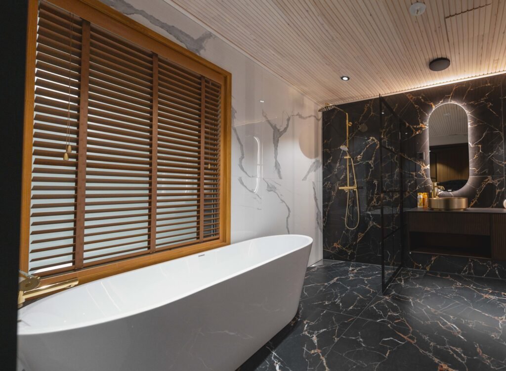 A freestanding bath beside a shuttered window, with a marble bathroom beyond
