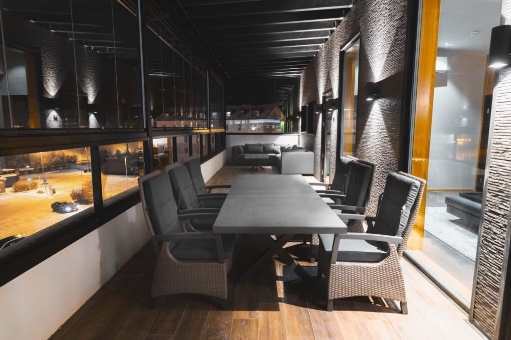 A long enclosed terrace with a dining table and armchairs, looking out over the city at night